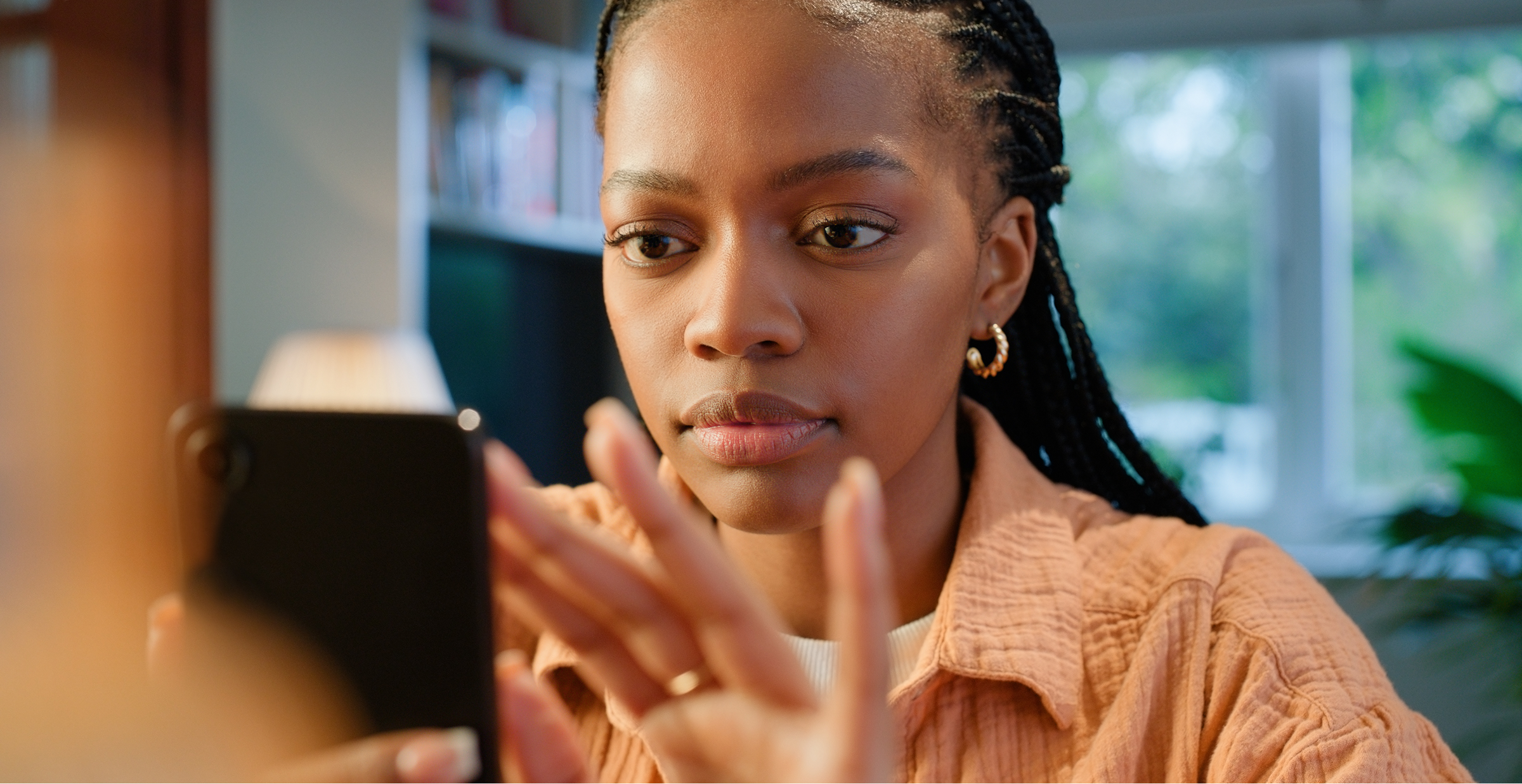 Person focused on using a smartphone in a warmly lit indoor setting with bookshelf and plants.